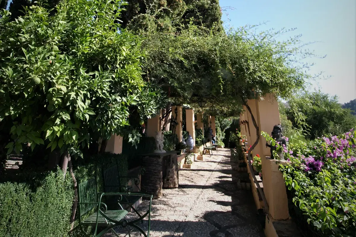 A shaded stone walkway under a vine pergola with tall cypress trees, orange bushes and flowering shrubs in a Granada carmen garden