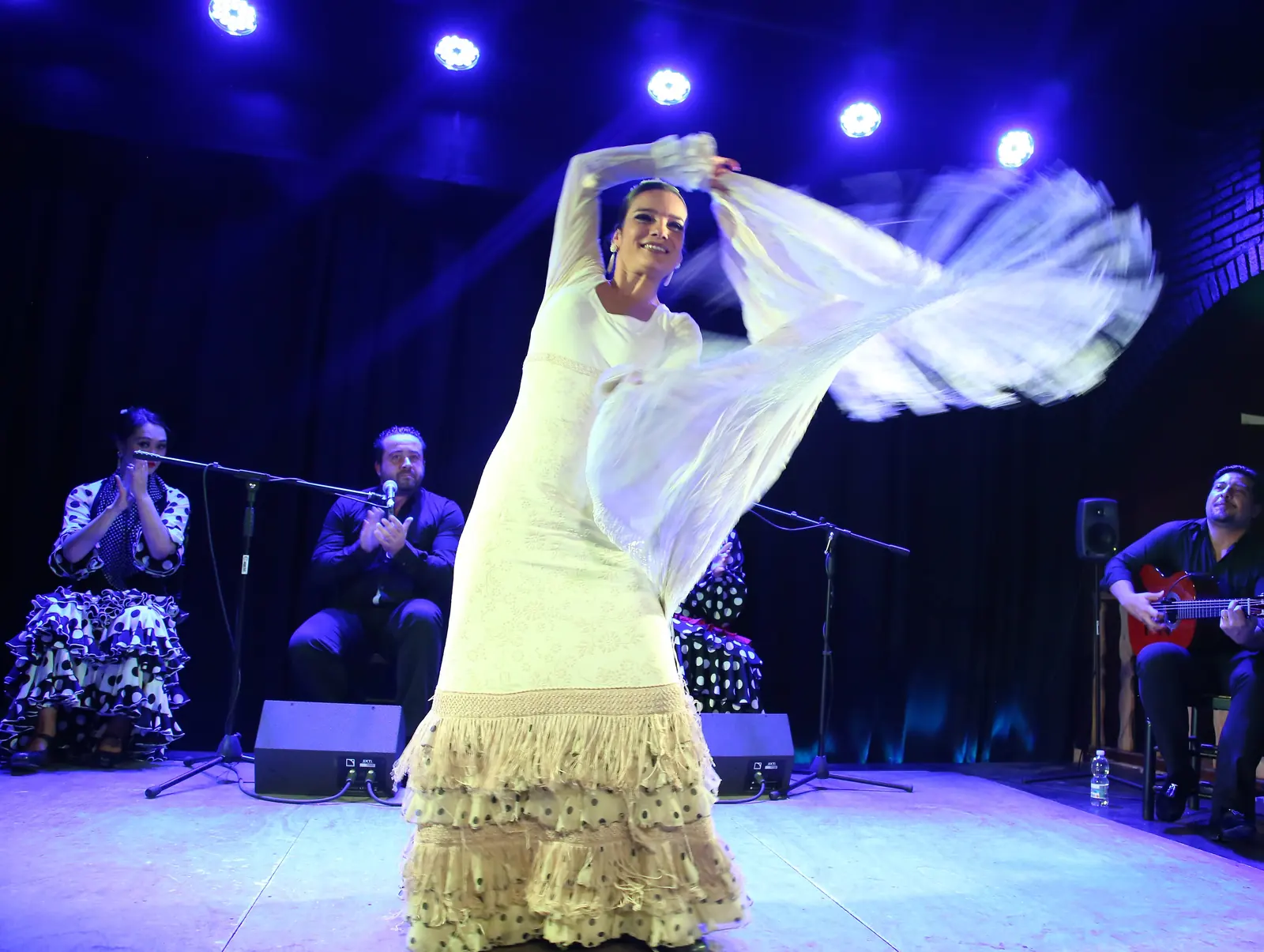 A flamenco dancer in a white dress and fringed shawl mid-turn on a blue-lit stage, with a guitarist and clapping artists seated behind her