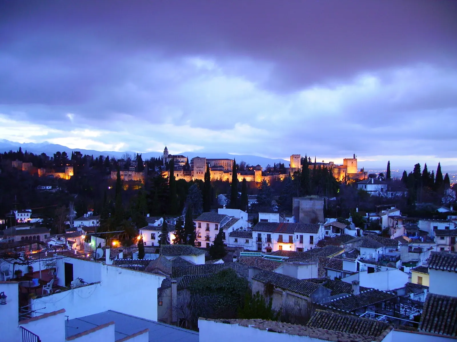 The lit Alhambra on its hill at dusk above the rooftops of the Albaicín, under a blue and violet sky