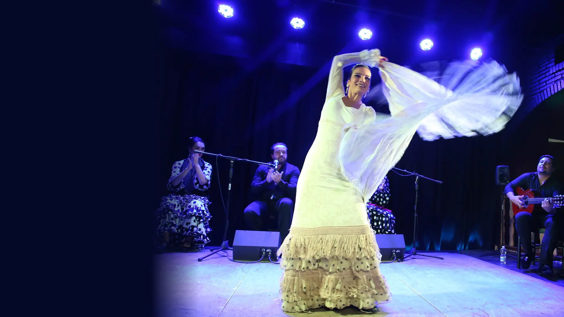 A flamenco dancer in a white dress and fringed shawl mid-turn on a blue-lit stage, with a guitarist and clapping artists seated behind her
