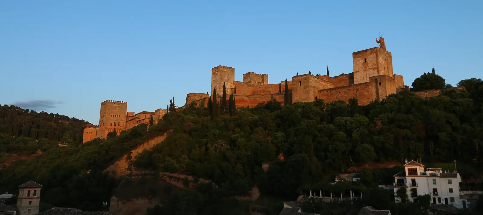 The red walls of the Alhambra on its wooded hill at sunset, seen from the Albaicín, with white houses at the lower right