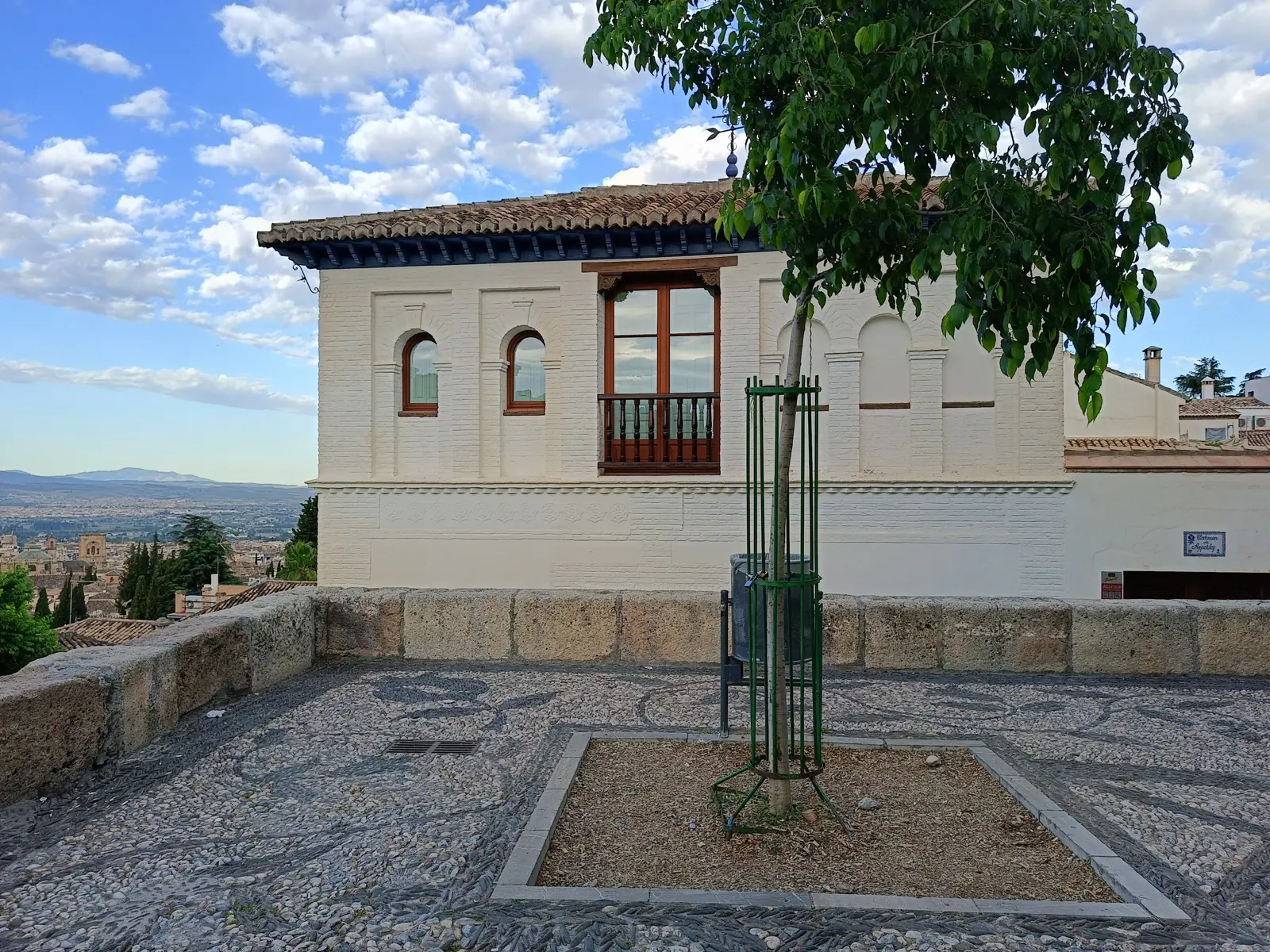 A white house with arched windows and a balcony beside a paved viewpoint terrace in the Albaicín, with the city far below